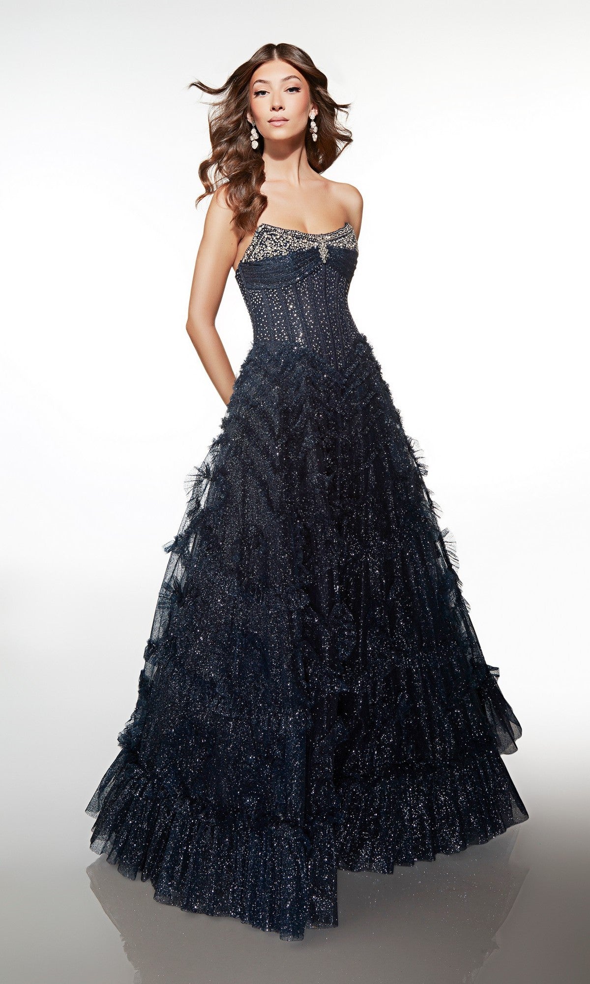 A woman poses in the Alyce 62096 long formal dress, showcasing a strapless embellished corset bodice and full ruffle-tiered tulle skirt in sparkling dark blue against a plain, light background.